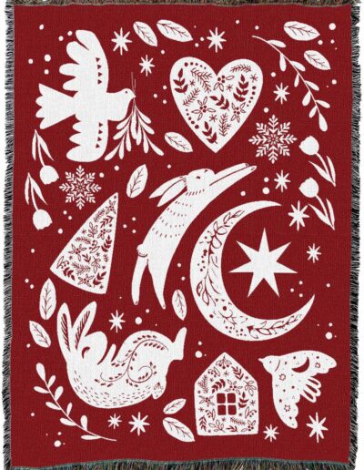 A red woven blanket patterned with white illustrations of rabbits, a dove, a heart, a crescent moon, a house, and various floral and snowflake motifs.