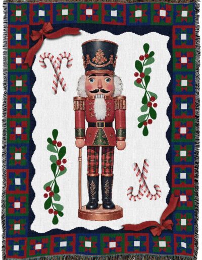Woven blanket featuring a central nutcracker surrounded by candy canes and holly, framed by a red, green, and blue geometric border with red ribbon accents at the corners.