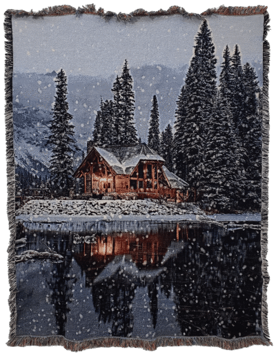 Woven throw blanket featuring a snow-covered log cabin illuminated at night, surrounded by pine trees and reflected in a dark lake.